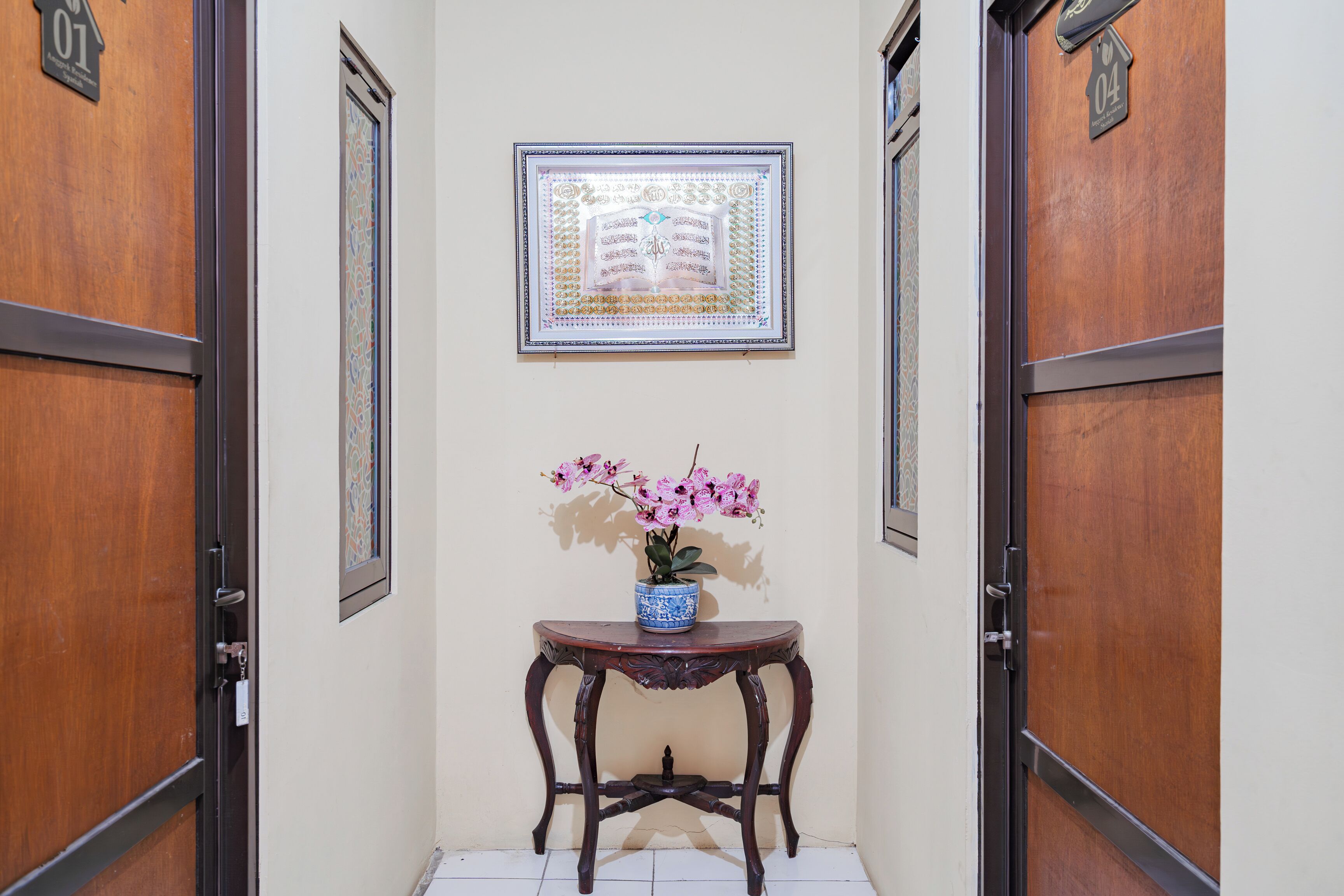 A hallway with wooden doors, a framed artwork above a table with a vase of flowers, and a tiled floor.