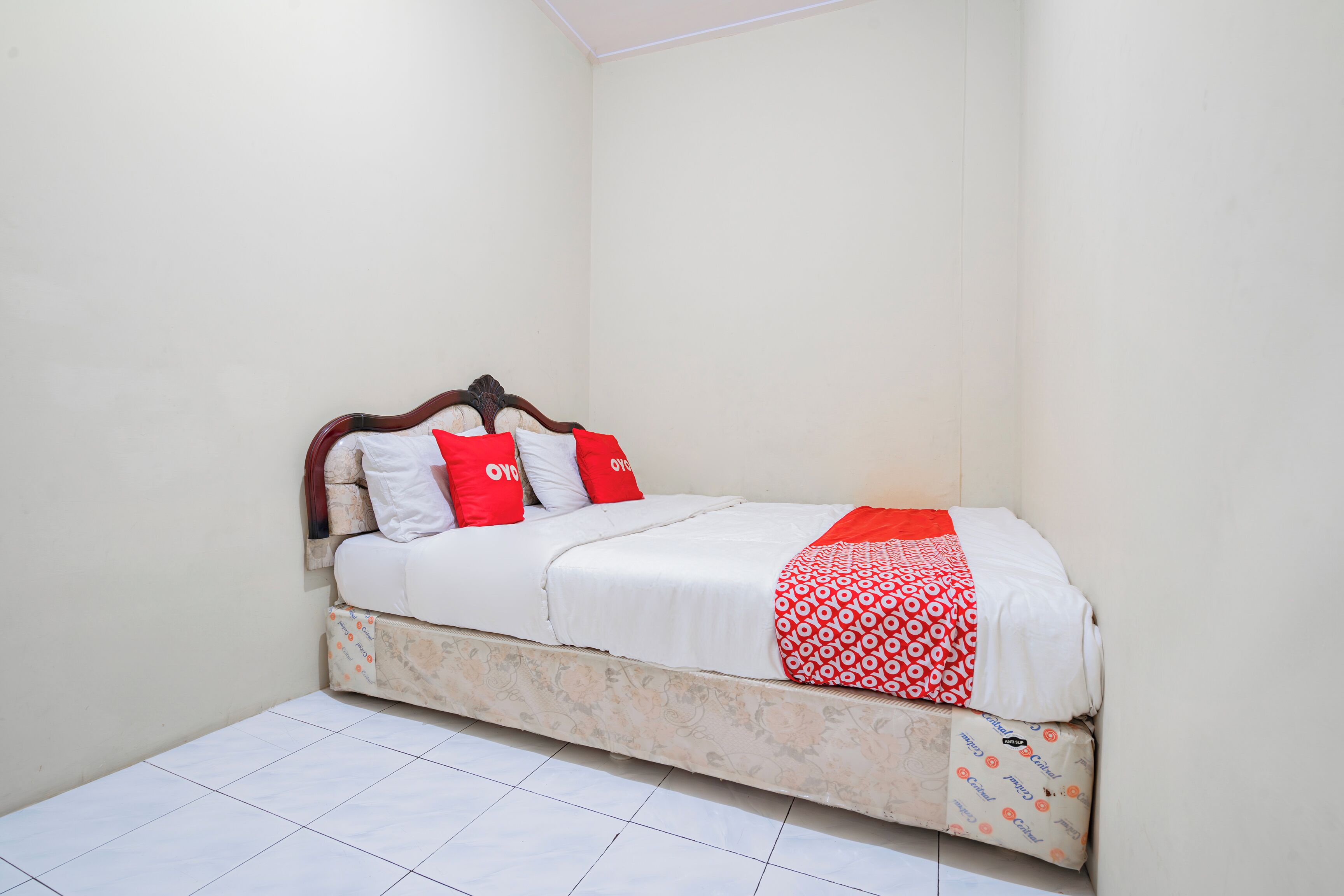 A small, neatly made bed with a patterned blanket and red pillows in a corner room with white walls and tiled floor.