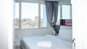 A modern bedroom with a large bed, a view of trees outside, and a window with curtains. - Vision condo (Pattaya)