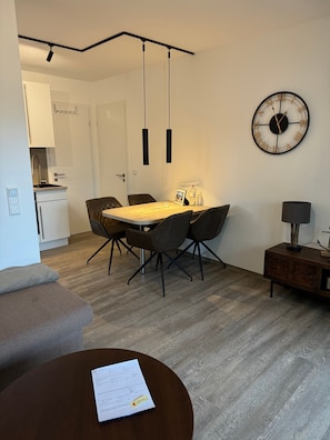 A modern living room with a dining area, a kitchen in the background, and a wall clock. - Apartment 'Residenz Lindengarten - Wohnung 16' with Private Terrace, Balcony and Wi-Fi (Kühlungsborn)