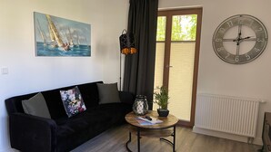 Una sala de estar con un sofá negro, una mesa redonda con una planta, un reloj y un cuadro de veleros.