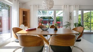 Un espace repas comprenant une table en bois, des chaises, un luminaire suspendu et une vue sur l’extérieur grâce à de grandes fenêtres.