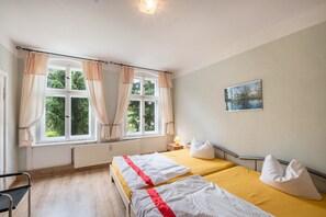 A hotel room with two beds, a window with curtains, a radiator, a bedside table with a lamp, and a painting on the wall.