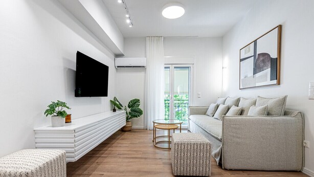 A modern living room with a flat-screen TV mounted on the wall, a grey sofa, a white console table with plants, and a wooden floor. - Phaedrus Living: Prosperity Residences Paradise (Athina)