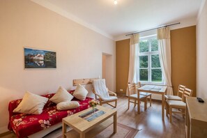Une pièce comprenant un canapé à motifs floraux rouges, une table basse en bois, une table à manger en bois avec des chaises, une fenêtre avec des rideaux et un tableau accroché au mur.