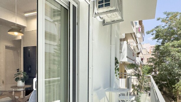 Uma varanda com móveis brancos, uma porta de vidro e vista para a cidade.