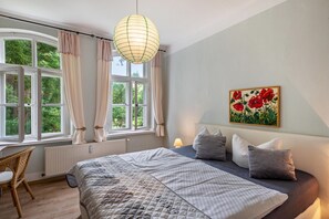 Une chambre avec un grand lit, une fenêtre avec des rideaux, un radiateur, un fauteuil en osier et un tableau au mur.