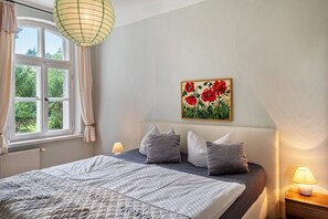 Une chambre à coucher avec un lit, une fenêtre avec des rideaux, un tableau représentant des fleurs rouges et une lampe de chevet.