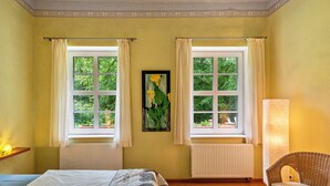 Une pièce avec deux fenêtres, un lit, une chaise et un radiateur.