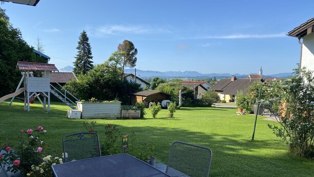 Een achtertuin met een patio tafel en stoelen, een speeltoestel voor kinderen, en een uitzicht op huizen en bergen in de verte.