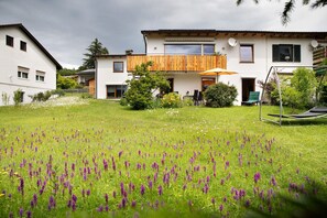 A two-story house with a balcony, a lawn with purple flowers, and a swing set.