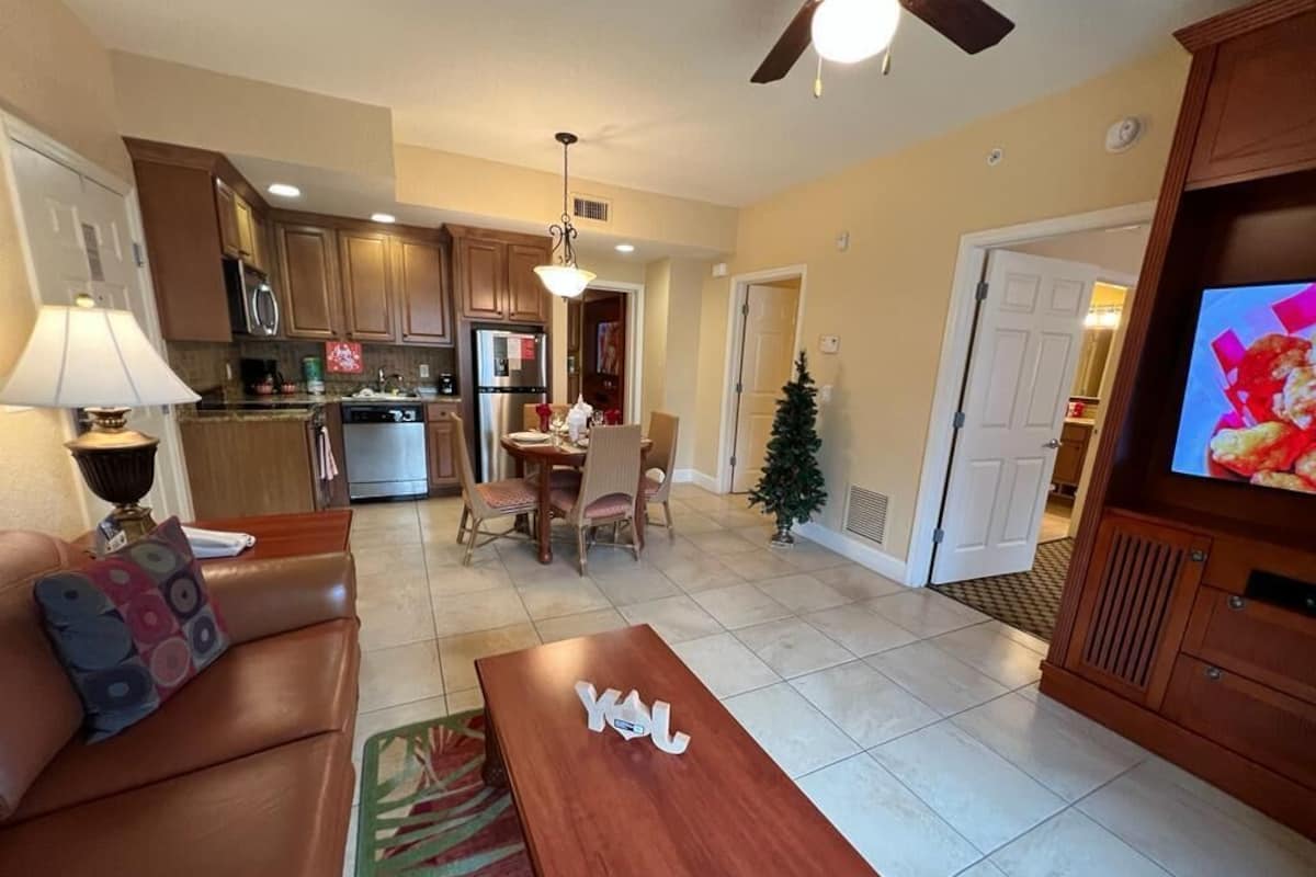 A living room with a sofa, coffee table, and a TV. There is a dining area with a table and chairs. The kitchen is visible with cabinets and appliances.