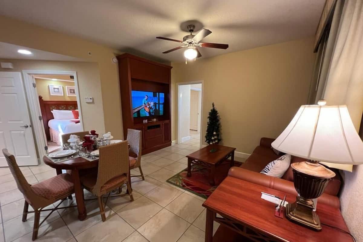 A hotel room with a dining area, a TV, a ceiling fan, and a lamp.
