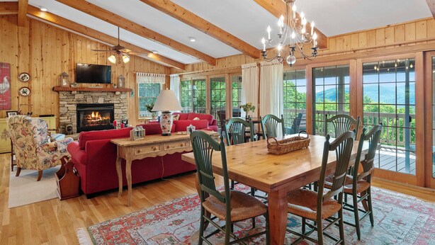 A spacious living room with a fireplace, wooden beams, a dining area with chairs, and large windows offering a view of the outdoors.