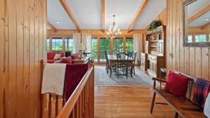 A spacious wooden interior with a dining area, a seating area with red cushions, and a large window offering a view of greenery.