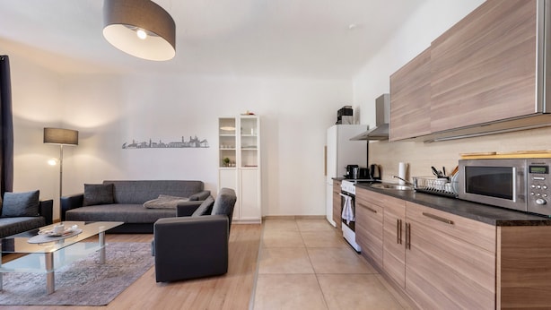 Un soggiorno moderno con divano, tavolino e televisore a schermo piatto. Adiacente, una cucina con forno a microonde, forno e mobili in legno.