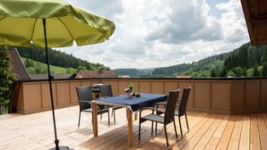 A wooden deck with a table and chairs, a green umbrella, and a view of a forested hill.