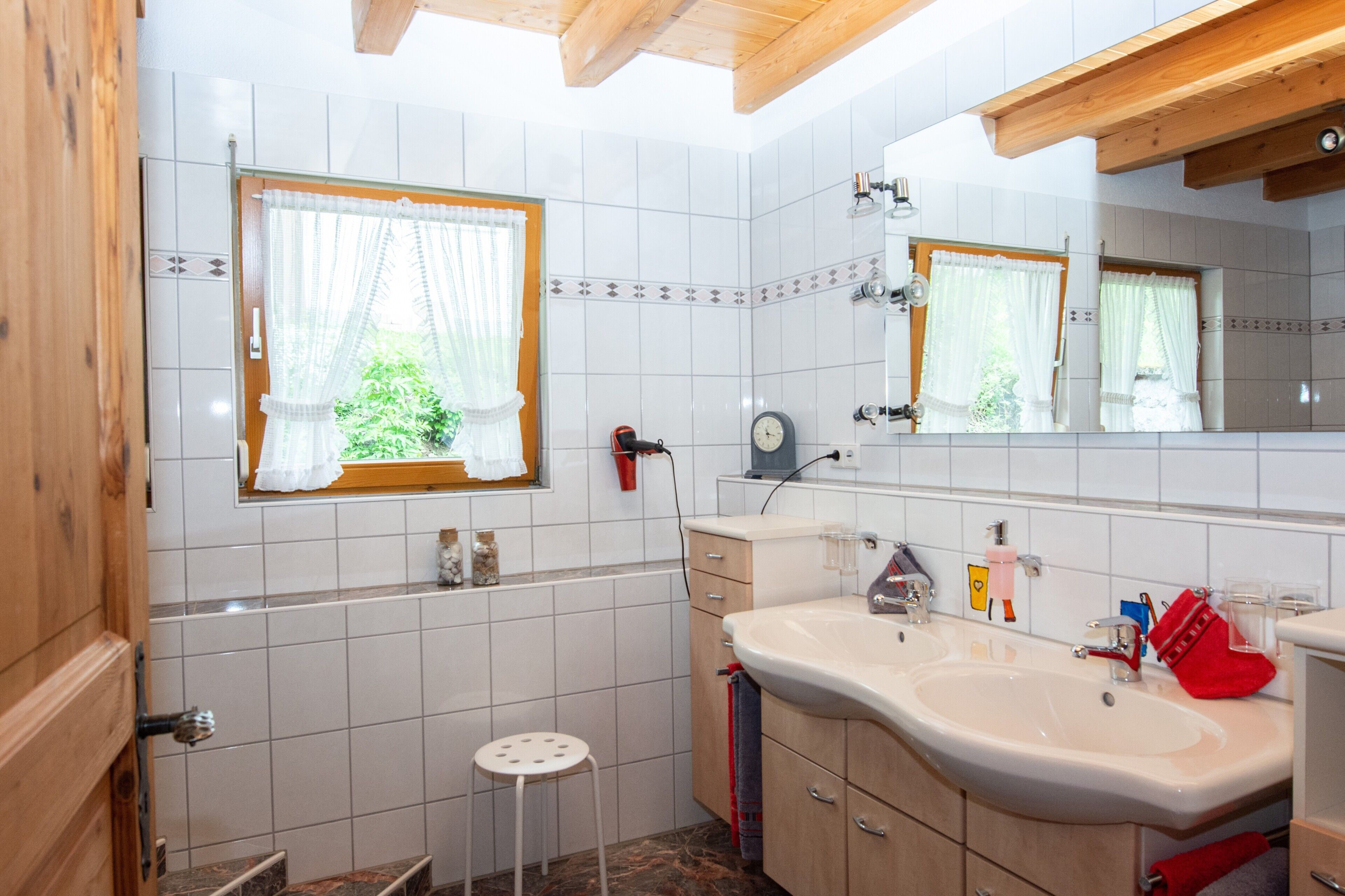 A bathroom with a large mirror, a sink, a window with curtains, and a wooden ceiling.