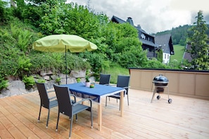 A wooden deck with a table and chairs, a grill, and a view of a house in the background. - Apartment 'Birgit' with Mountain View, Private Terrace and Wi-Fi (Schonach im Schwarzwald)