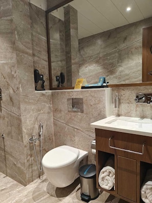 A modern bathroom with a marble floor, a white toilet, a shower area, and a wooden vanity with a sink. - Kings Kraft Tremezzo Inn Somnath (Veraval)