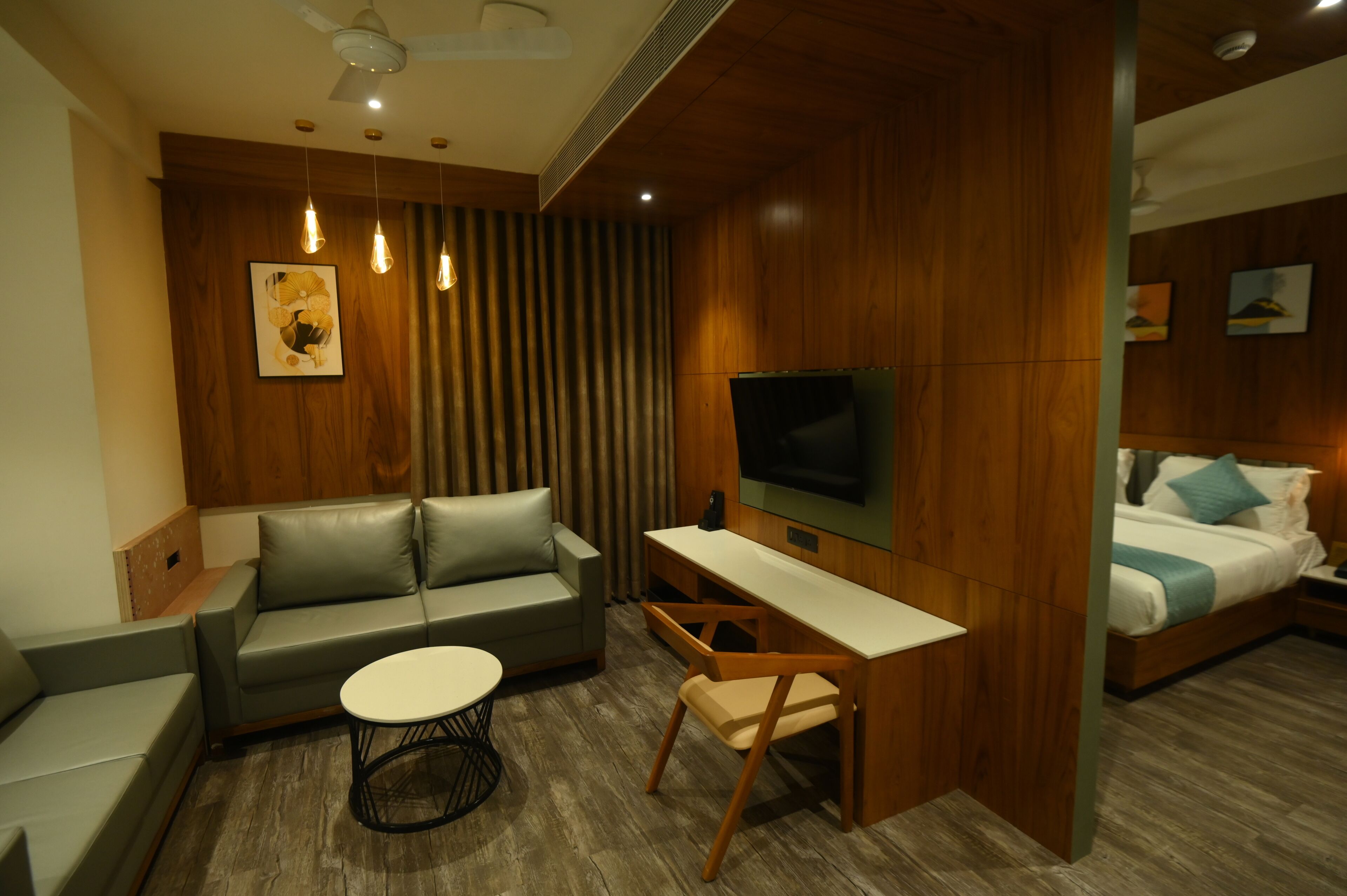 A modern hotel room with a sofa, a small round table, a desk with a chair, a TV, and a bed with a blue pillow.