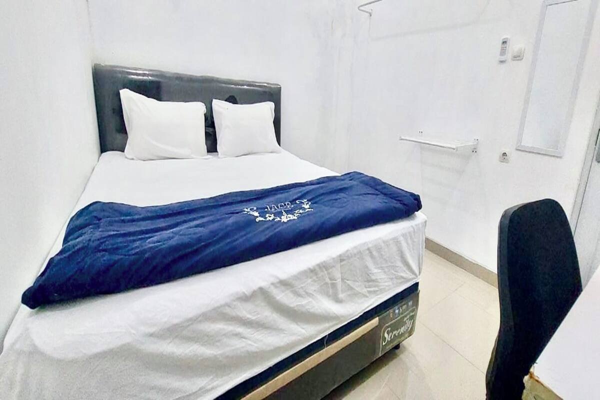 Una cama individual con una colcha blanca y azul, un cabecero oscuro, y una pared blanca con una puerta y un interruptor de luz.