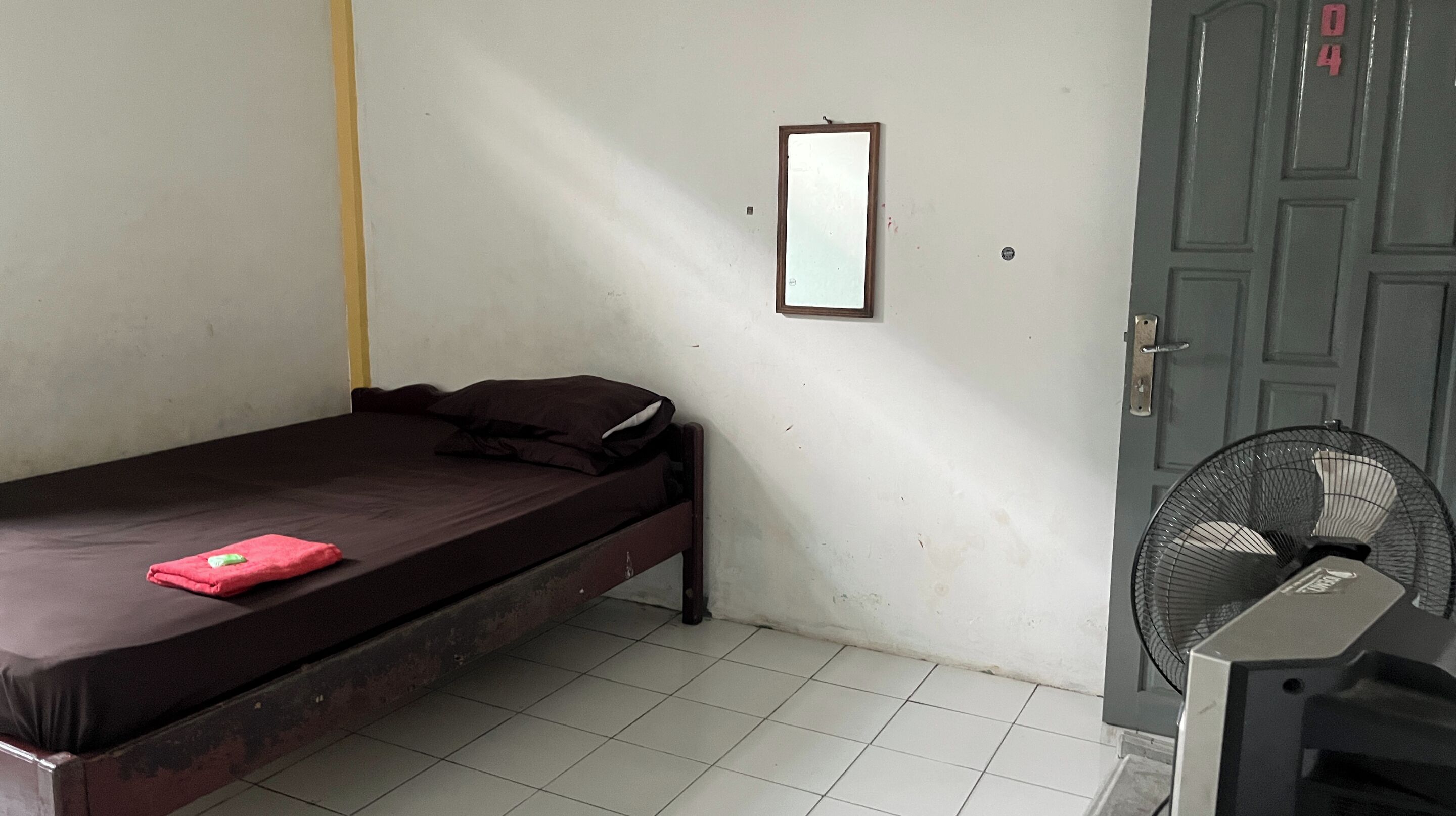 Ein kleines Zimmer mit einem Einzelbett, einem hölzernen Bettgestell, einem kleinen Spiegel an der Wand, einem Ventilator und einer Tür mit einer Nummer.
