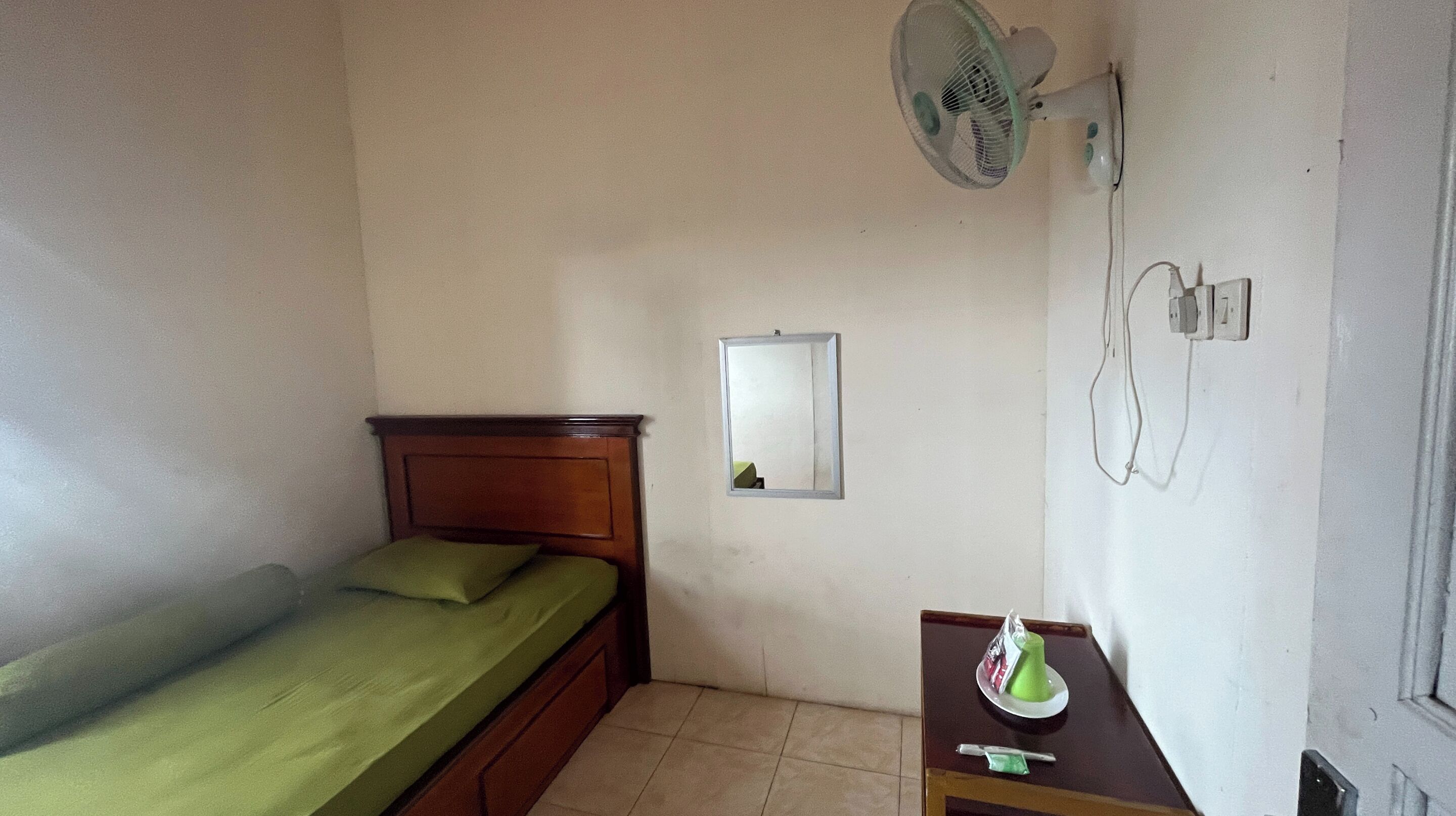 Een kleine, eenvoudige kamer met een enkel bed, een ventilator aan de muur, een spiegel en een klein tafeltje met een groen voorwerp erop.