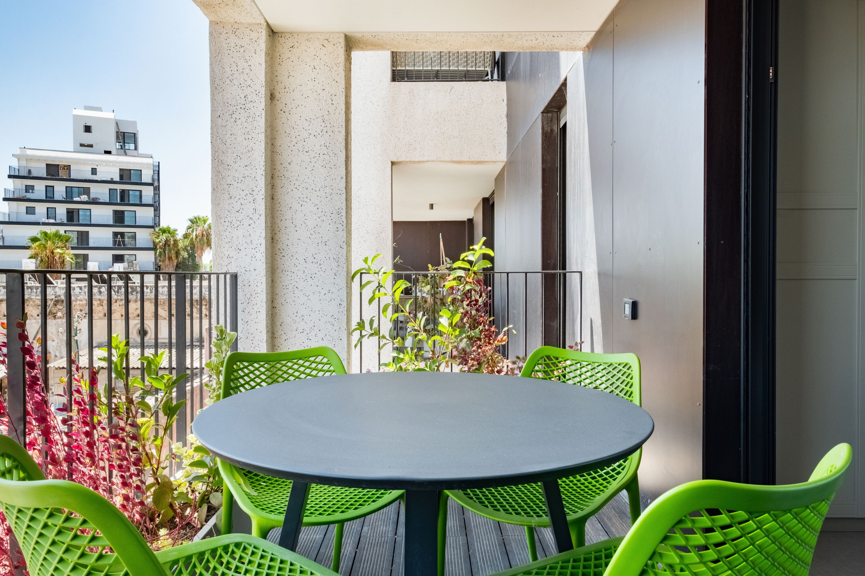 A balcony with a round table and green chairs, surrounded by plants and a view of a building.