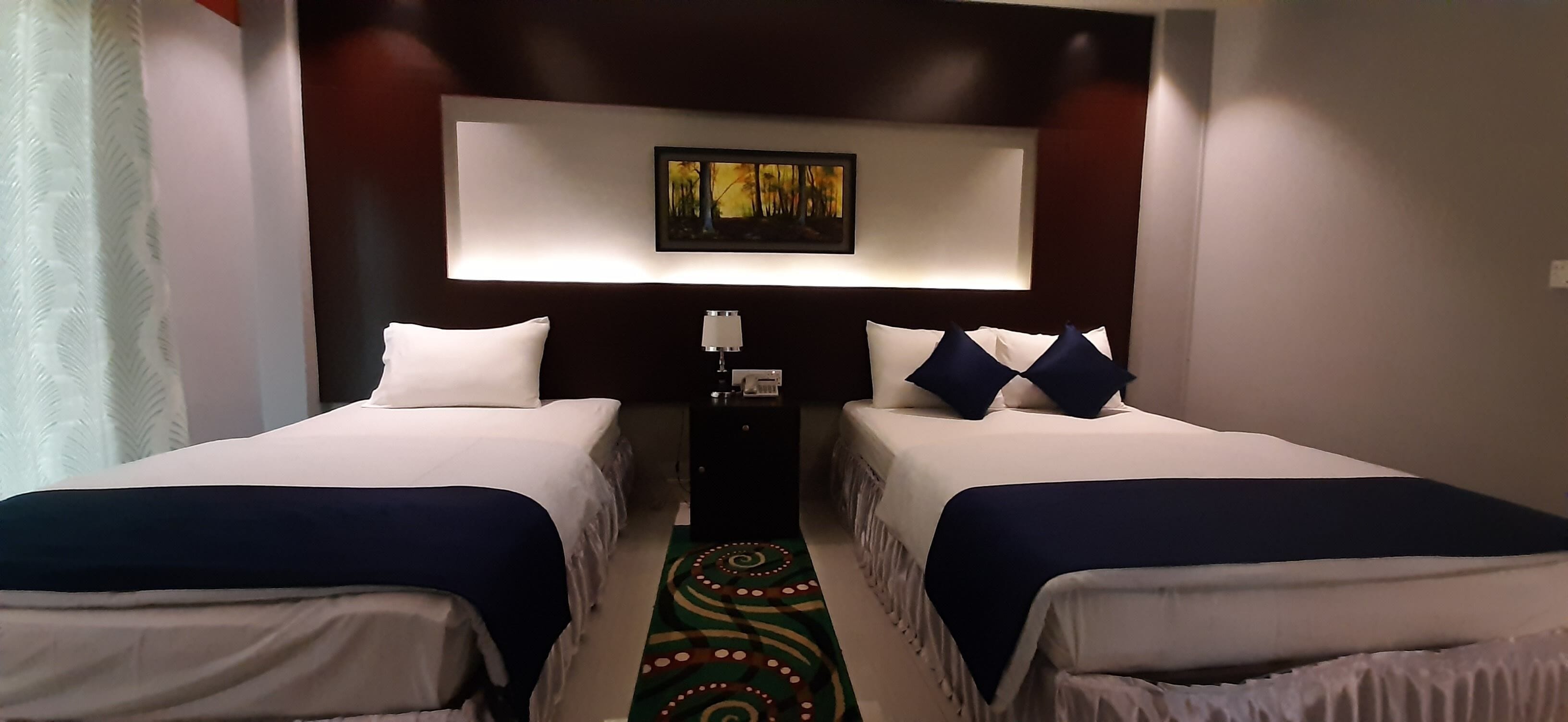 A hotel room with two single beds, a nightstand with a lamp, a painting on the wall, and a patterned rug.