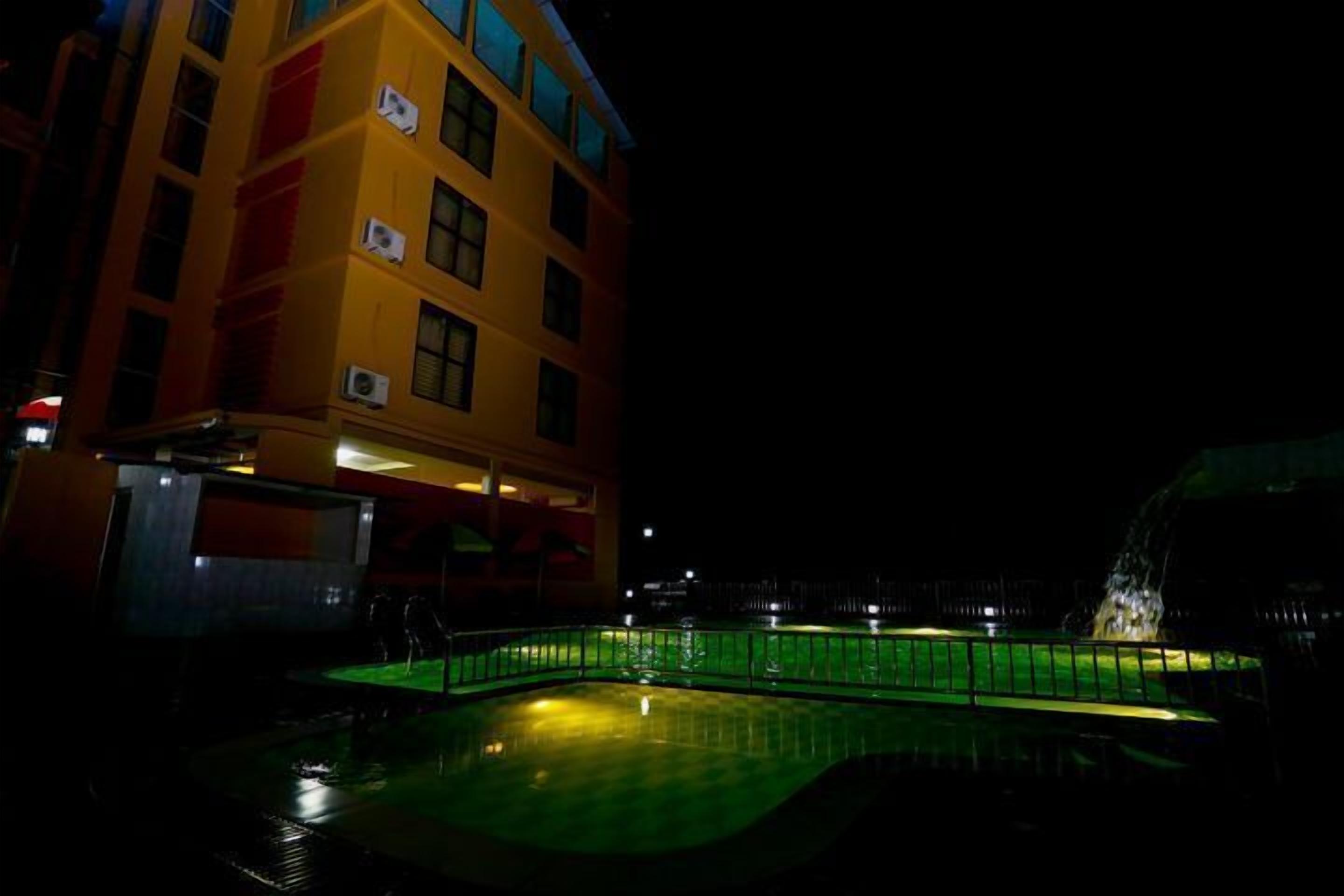 Un edificio con varios pisos y balcones, una piscina con plataforma iluminada en verde y una fuente de agua al fondo.