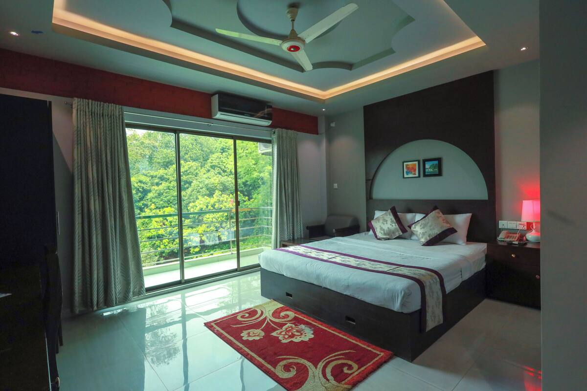 A modern bedroom with a large bed, a ceiling fan, a red rug, and a view of greenery through the window.