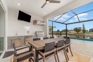 A modern outdoor kitchen with a large dining table, chairs, a grill, and a TV mounted on the wall.