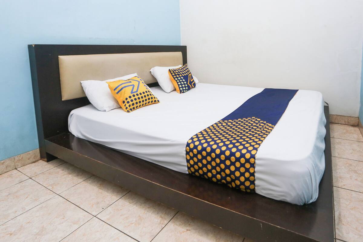 Una cama con colchón blanco, dos almohadas, una manta de tonos azules y amarillos, y un cabecero de madera oscura.