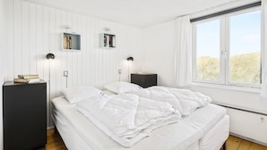 Ein Schlafzimmer mit einem Bett, zwei Nachttischen, zwei Wandregalen mit Büchern, einem Fenster mit Blick ins Freie und einer Holzwanddecke.