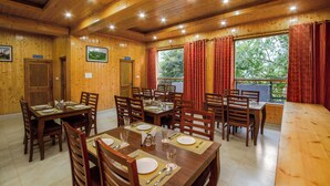 A dining area with wooden walls and ceiling, multiple tables set for dining, and a view of trees through large windows. - Foxoso Cherry Hotel Manali (Manali)