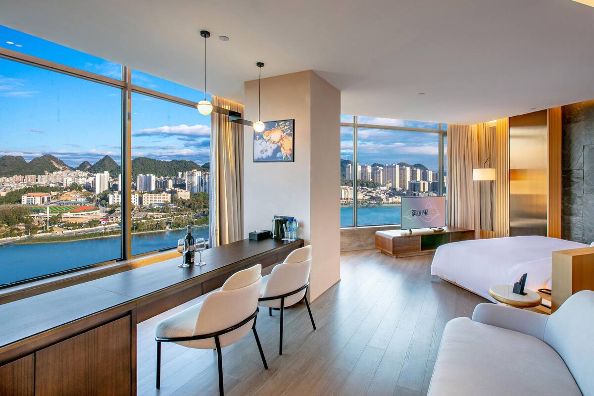 Une chambre d’hôtel moderne offrant une vue imprenable sur la ville, avec un bureau et des chaises, un lit et un petit coin salon.
