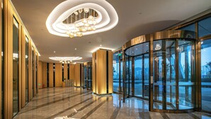 Un hall d’hôtel moderne avec une entrée en verre incurvé, un lustre central et un sol en marbre poli.