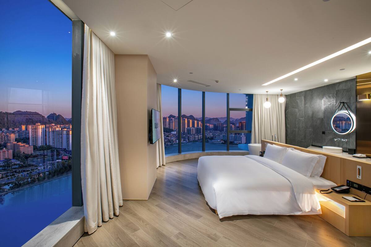 Une chambre d’hôtel moderne dotée d’un grand lit, d’une salle de bain avec un miroir et offrant une vue sur la ville grâce à de grandes fenêtres.
