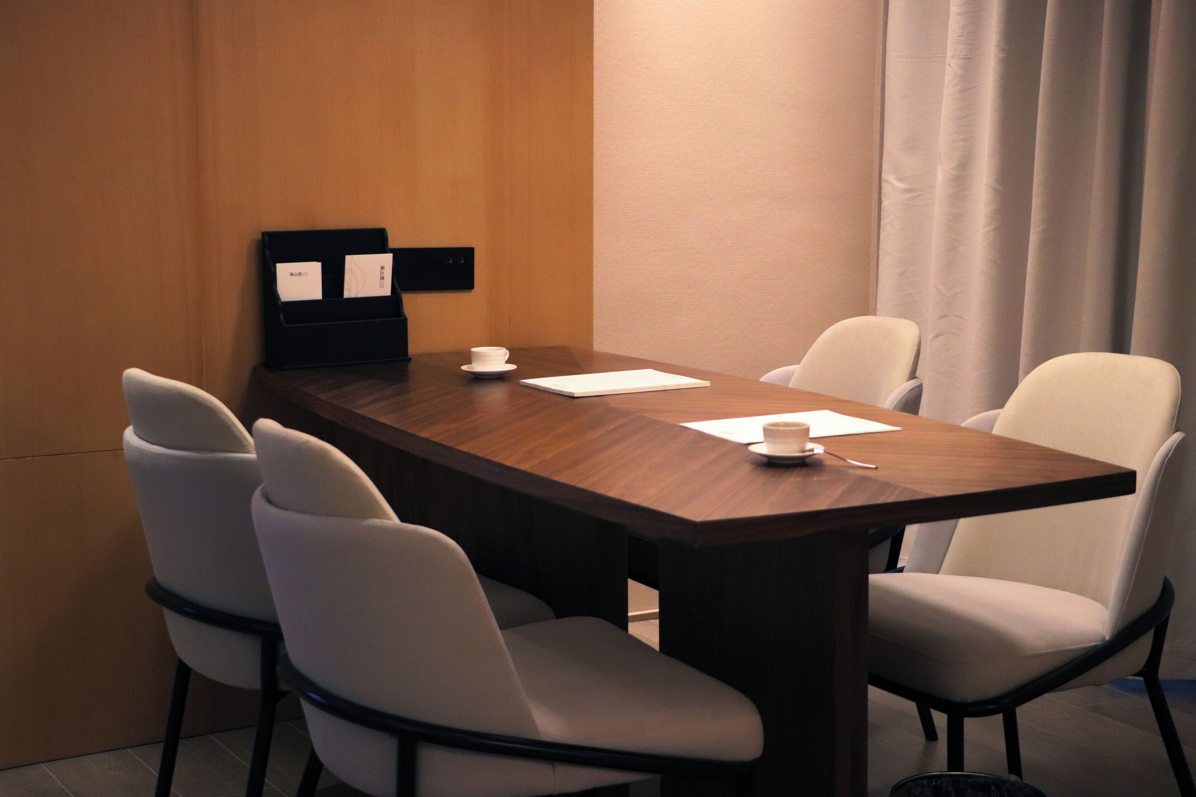 Un salón de conferencias con una mesa de madera, cuatro sillas blancas, una pizarra con papeles y una taza blanca sobre la mesa.