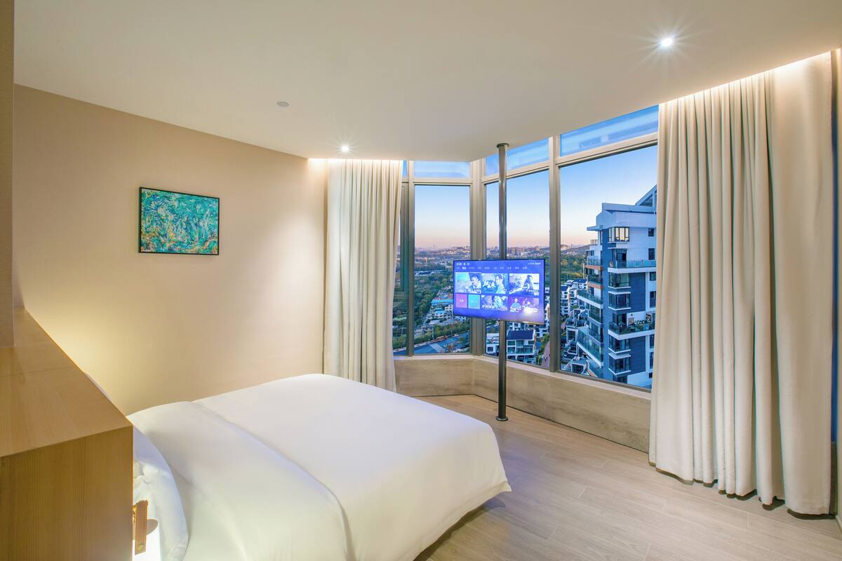 Une chambre d’hôtel avec un grand lit, un téléviseur fixé au mur et une vue sur le paysage urbain grâce à de grandes fenêtres.