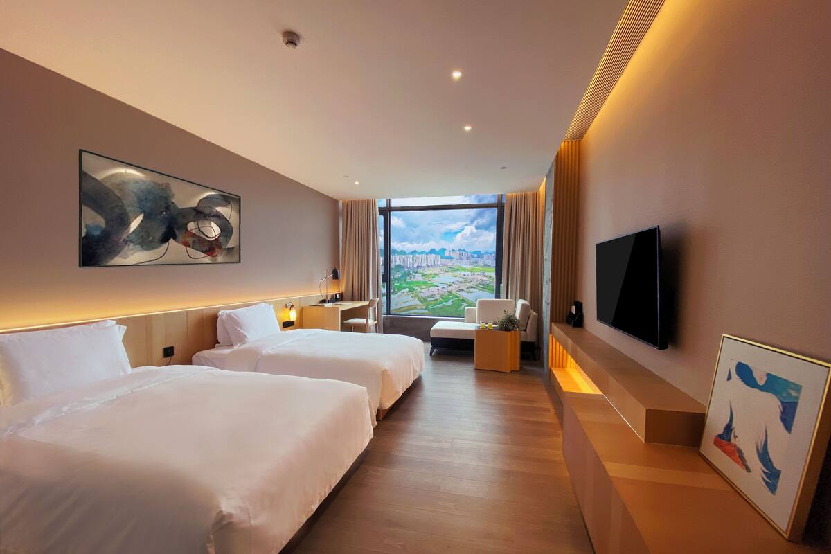 Une chambre d’hôtel avec deux lits, un bureau, une chaise, une télévision et une grande fenêtre donnant sur la ville.