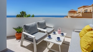 Ein Balkon mit einer weißen Couch, zwei grauen Sesseln, einem kleinen Tisch mit blauen Tassen und einem Blumentopf.