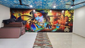 A room with a mural of a deity playing a flute, a bed with plaid bedding, and a ceiling fan. - Shree Keshav Haweli (Sikar)