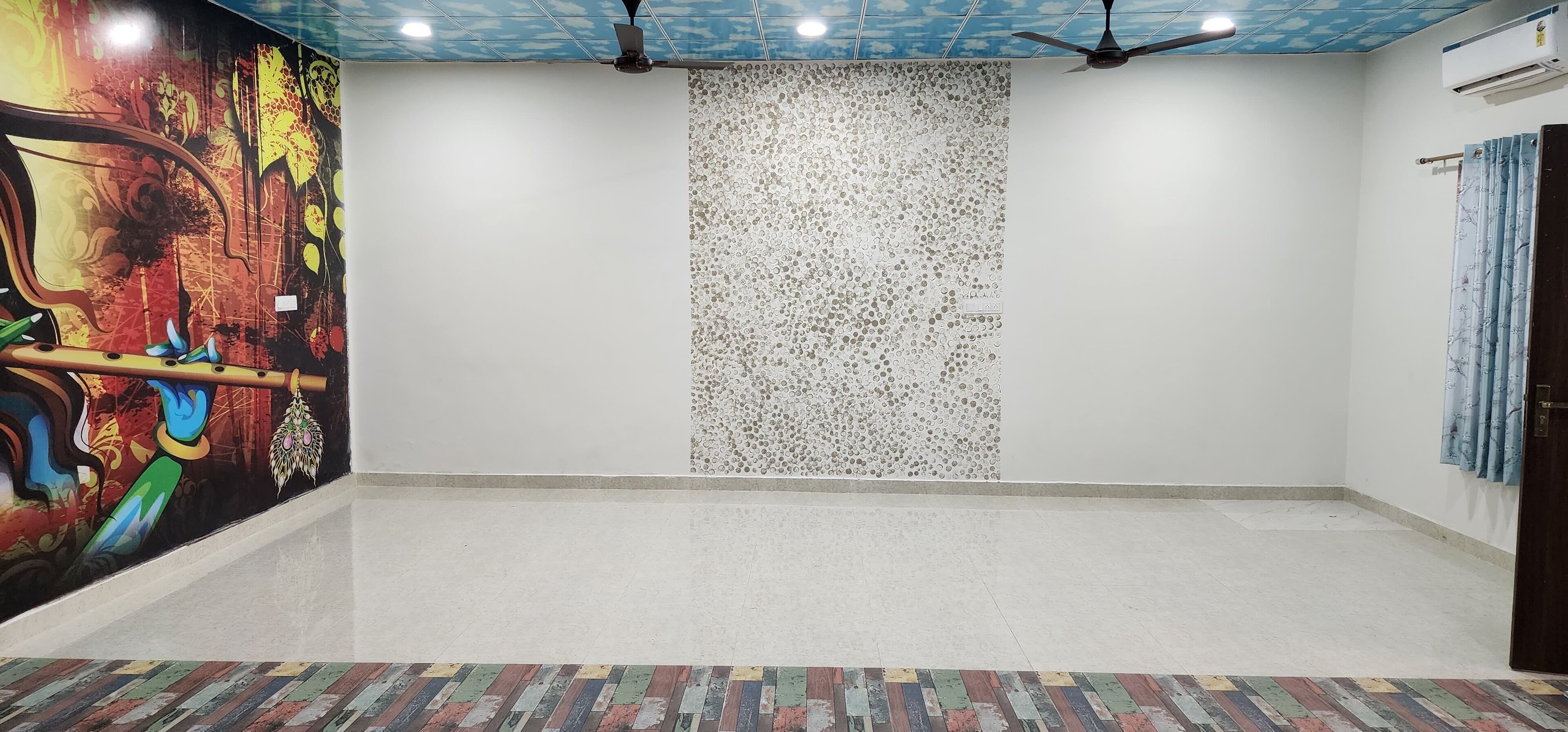 Una habitación amplia con un mural colorido en una pared, un separador decorativo y un ventilador de techo.