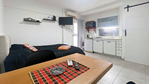 A compact living space with a bed, a small dining table, a kitchenette, and a TV mounted on the wall. - Cozy Studio in Palermo with Private Patio! (Buenos Aires)