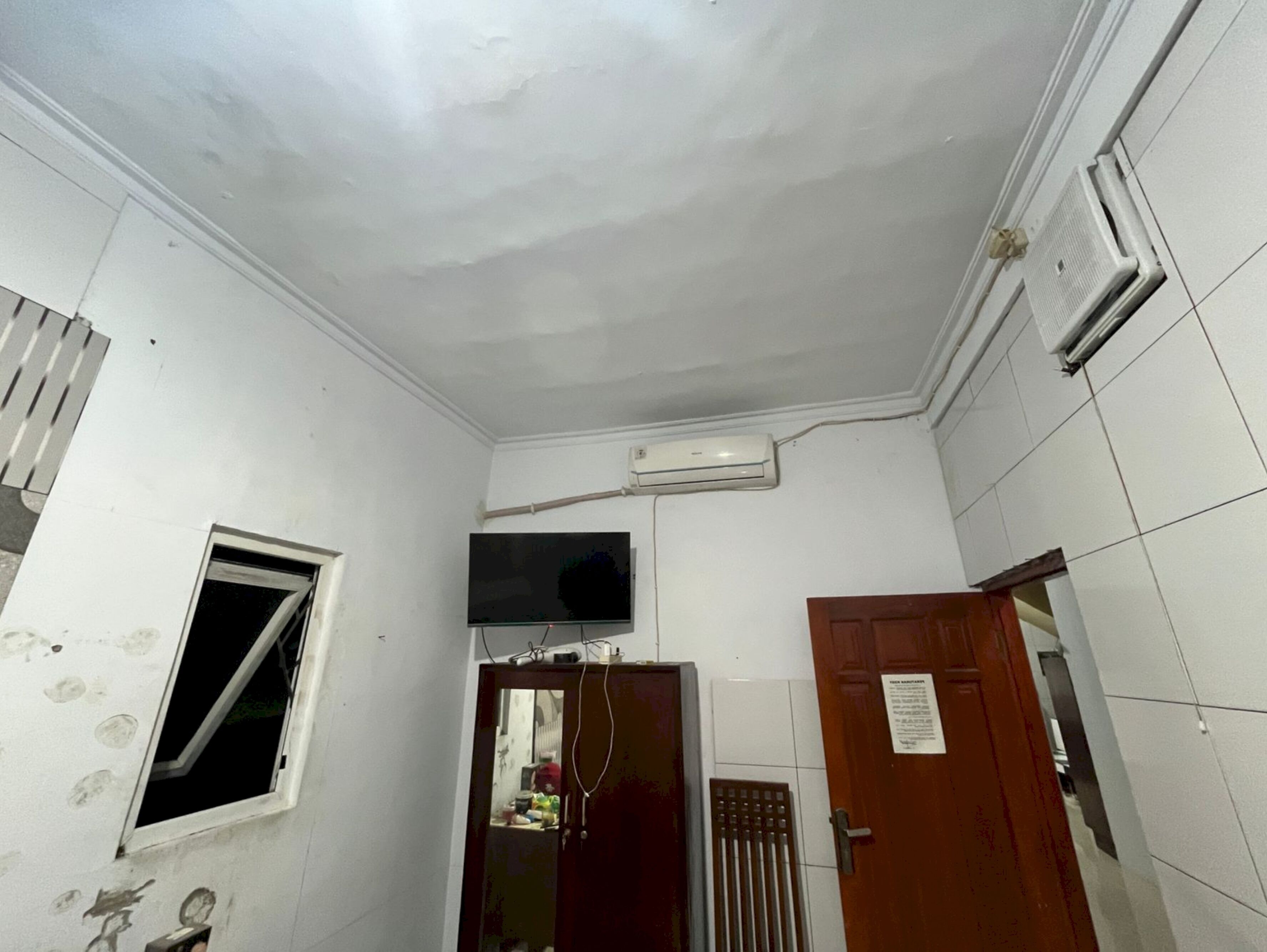 Una stanza piccola e poco illuminata, con pareti bianche, una finestra, una porta e un condizionatore d'aria fissato al muro.