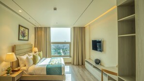 A hotel room with a bed, bedside lamps, a TV, a window with curtains, and a view of the city. - juSTa Residences - AIPL Joy Street Gurgaon (Gurugram)
