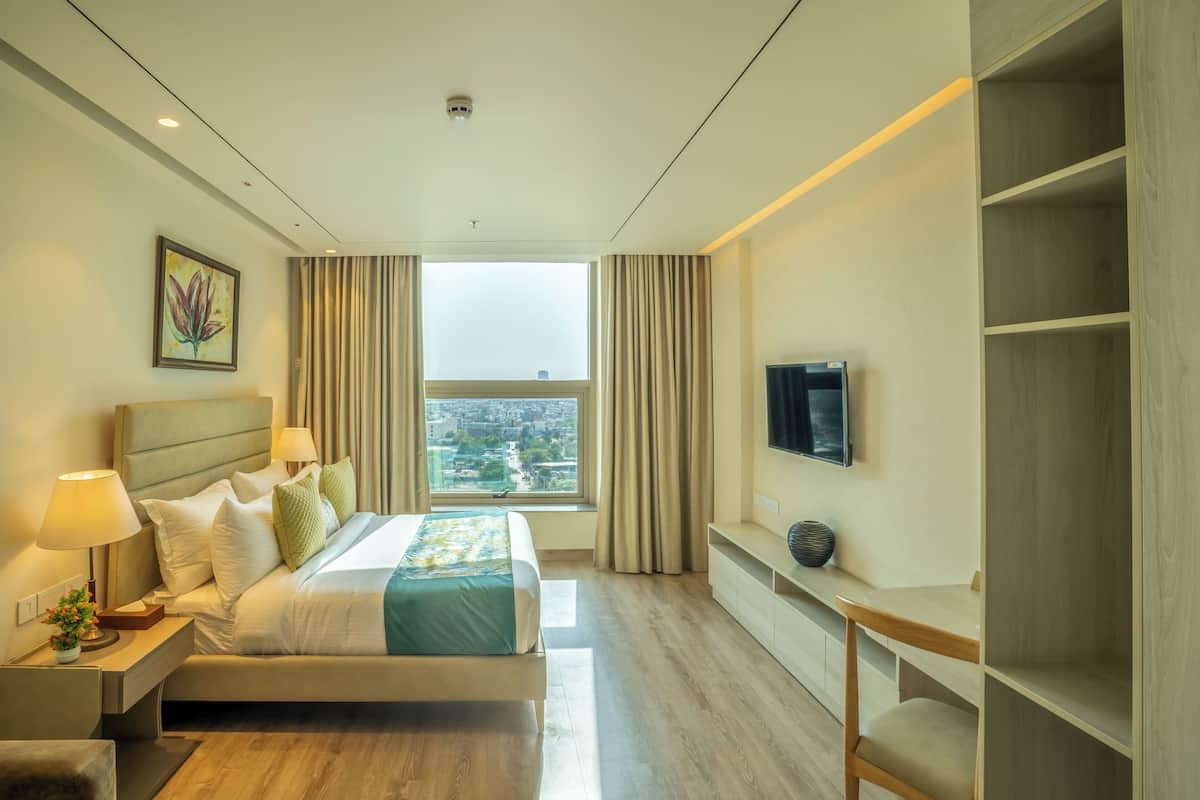 A hotel room with a bed, bedside lamps, a TV, a window with curtains, and a view of the city.