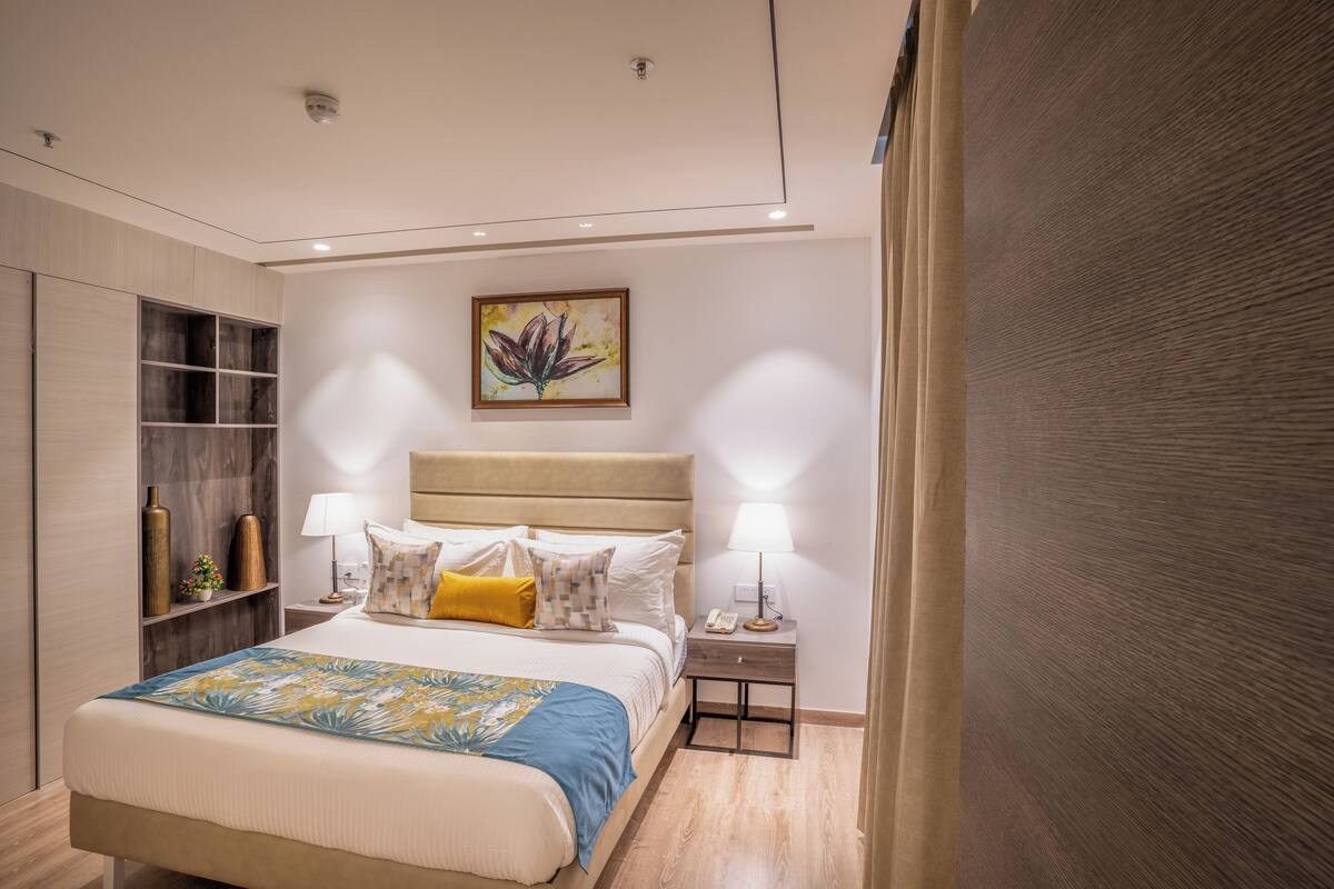 A modern bedroom with a large bed, bedside tables, a nightstand, a wall-mounted shelf, and a ceiling light fixture.
